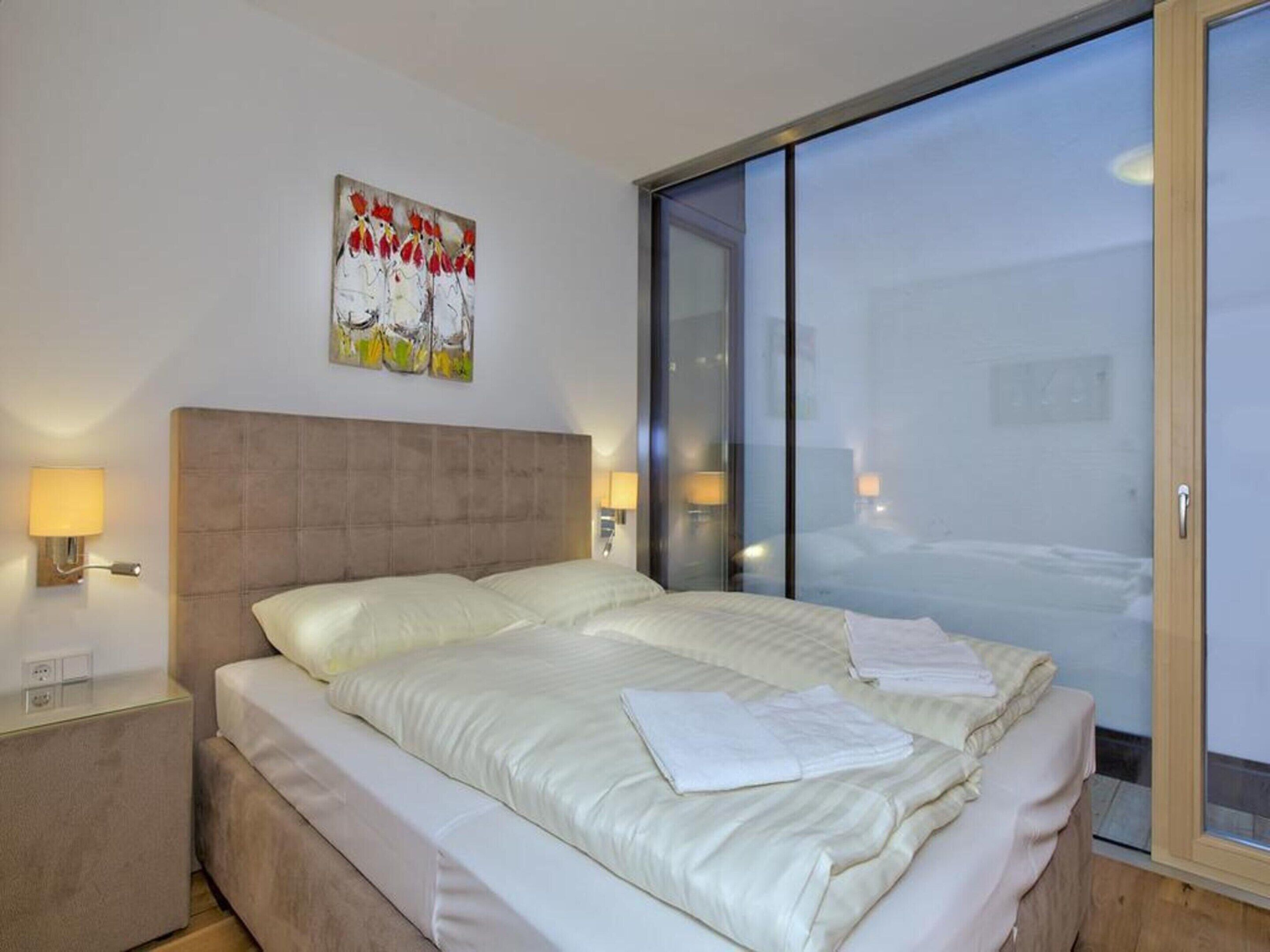 A hotel room with a bed, a nightstand, a wall-mounted lamp, a painting, and a large window.