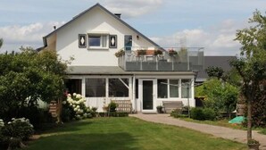 Une maison à deux étages dotée d’un balcon, d’une porte d’entrée et d’un petit jardin agrémenté de fleurs et d’un banc.