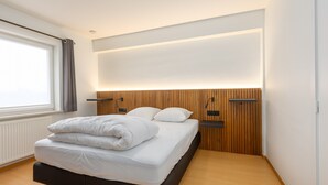 A modern bedroom with a bed, wooden headboard, bedside tables, and a window with curtains. - Cozy Apartment Near the Beach (Middelkerke)