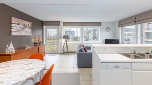 A modern kitchen with a built-in dishwasher, a dining area with a table and chairs, and a large window with a view of the city. - Cozy Apartment Near the Beach (Middelkerke)