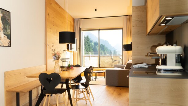 Eine moderne Küche mit Holzschränken, ein Essbereich mit einem Holztisch und -stühlen und ein großes Fenster mit Blick ins Freie.