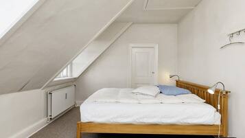 Ein Schlafzimmer mit geneigter Decke, einem Bett mit weißer Bettwäsche, einem Holz-Kopfteil, einer Nachttischlampe und einem Heizkörper.