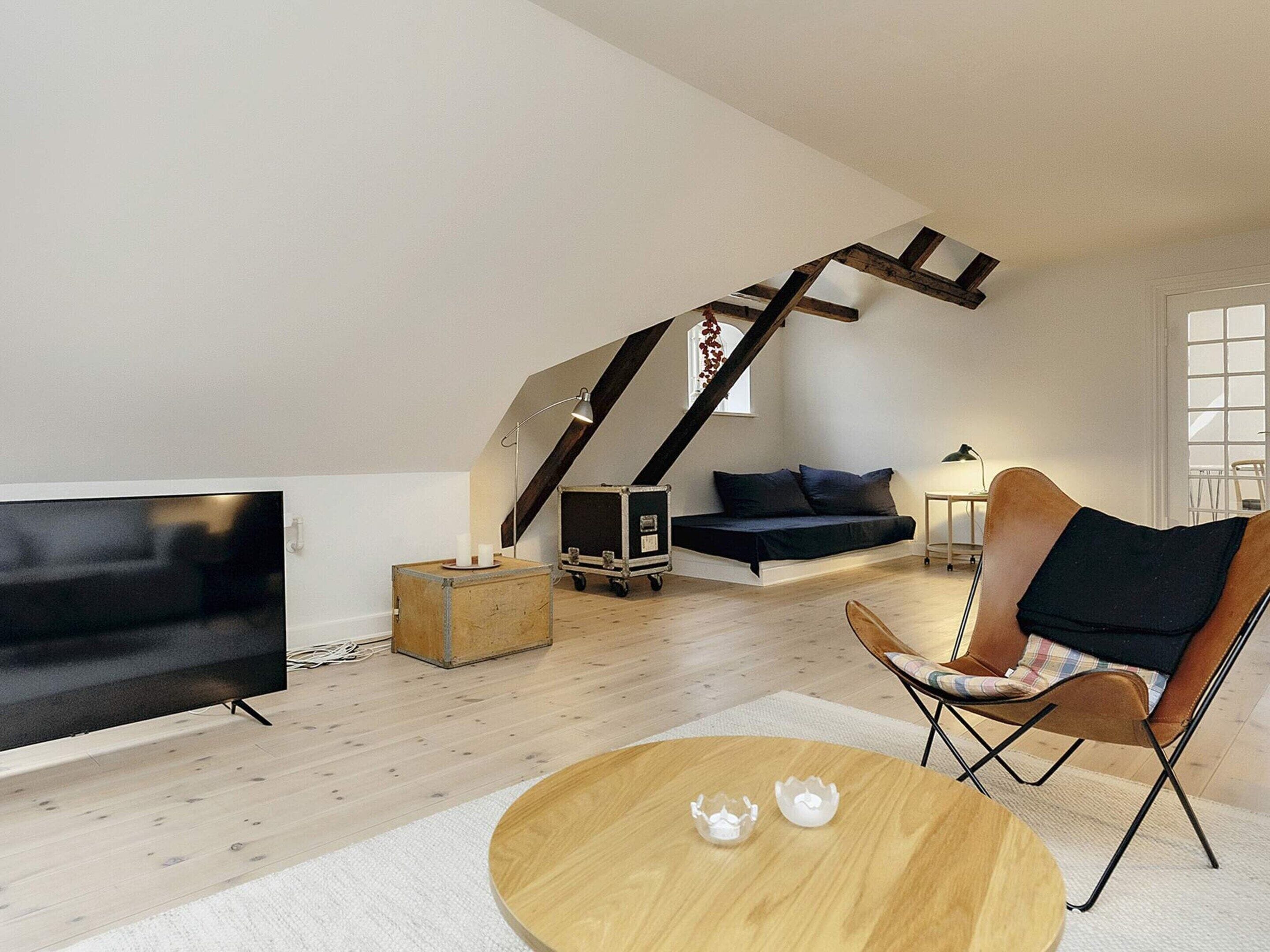 Een woonkamer in loft-stijl met een hoge plafond, een houten trap, een zwarte bank, een bruine leren fauteuil en een ronde houten salontafel.