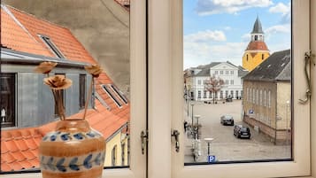 Blick aus dem Fenster auf einen Stadtplatz mit Gebäuden, einem Kirchturm und einem parkenden Auto.