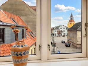 Une vue par la fenêtre sur une place de ville, avec des bâtiments, le clocher d’une église et une voiture garée.