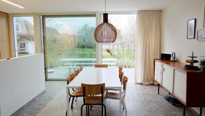A modern kitchen with a large dining table, chairs, a hanging light fixture, and a view through the glass door. - Apartment in Nieuwpoort by Beach (Koksijde)