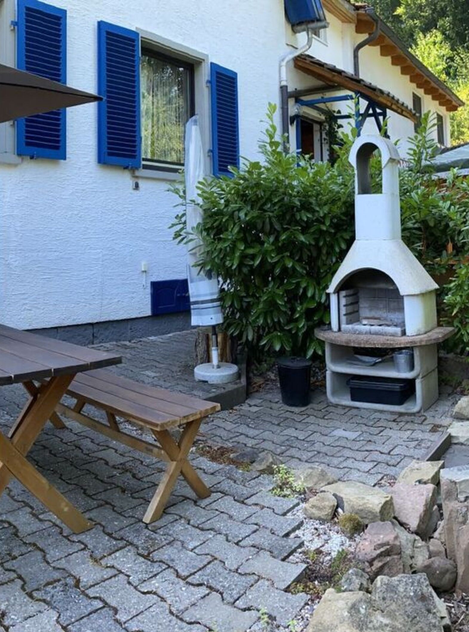 Un espace patio avec une table et des bancs en pierre, un petit four d’extérieur et un bâtiment blanc aux volets bleus.