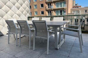 Un balcón con una mesa y sillas, suelo de baldosas y un edificio al fondo con varias ventanas y balcones.