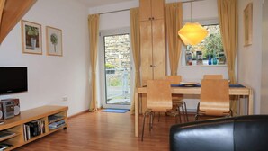 A modern living room with a flat-screen TV, wooden flooring, a dining area with a table and chairs, and a large window with curtains. - Holiday Apartment Riesling (Lorch)