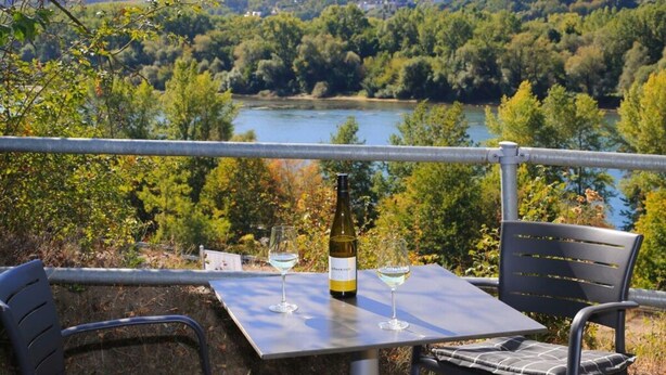 A table with two wine glasses and a bottle, overlooking a river and forested hills. - Holiday Apartment Riesling (Lorch)