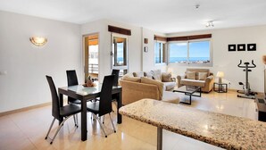 A modern living room with a dining area, a large window with a view of the sea, and a ceiling fan. - Belvilla by OYO Apartamento Sol y Paraaso I (Adeje)