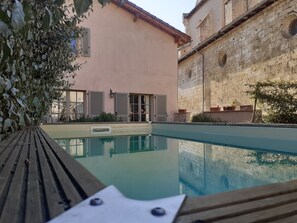A pool area with a wooden deck, a building with shuttered windows, and a stone wall. - Le Clos de Sarrali Maison de Charme (Sarrant)