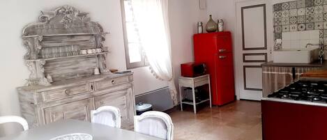 A dining area with a white table and chairs, a vintage-style wooden cabinet, a red refrigerator, and a kitchen area with tiled backsplash and a sink.