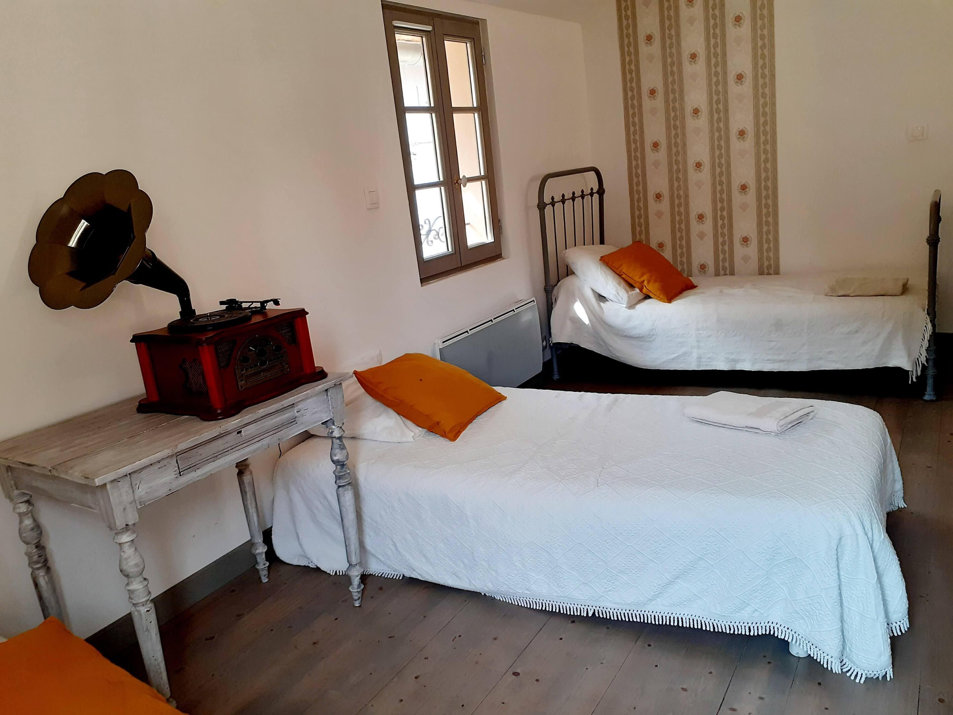 A room with two single beds, a wooden desk with a gramophone, a radiator, and a window with curtains.