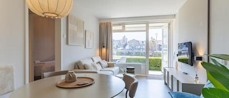 A modern living room with a dining area, a sofa set, a television mounted on the wall, and a large window offering a view of the city.