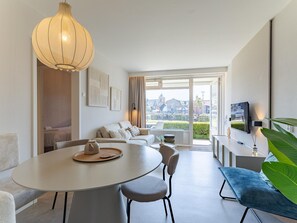 Un salon moderne comprenant un coin repas, un ensemble de canapés, un téléviseur fixé au mur et une grande fenêtre donnant sur la ville.