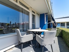 An outdoor seating area with a table and four wicker chairs, positioned next to a building with large windows and a blue accent wall.