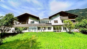 A two-story building with a balcony, surrounded by lush green grass and trees, set against a mountainous backdrop. - Spacious Apartment in Gortipohl With Garden (Sankt Gallenkirch)