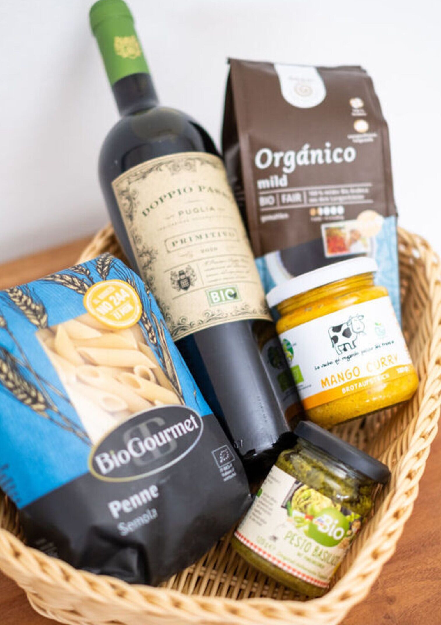 Una cesta con una botella de vino, una bolsa de café, una bolsa de pasta y frascos de pesto y curry de mango.