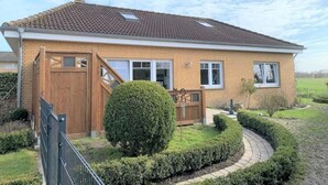 A single-story house with a tiled roof, a wooden front gate, and a well-maintained garden with a circular hedge and a small tree. - Ostfriesland to Huus (Hinte)