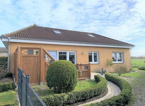 A single-story house with a tiled roof, a wooden front gate, and a well-maintained garden with a circular hedge and a small tree. - Ostfriesland to Huus (Hinte)
