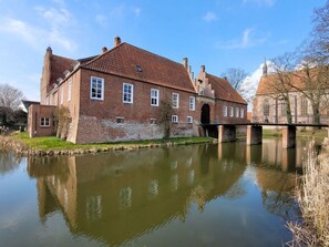 A historic brick building with a river in front, a bridge, and a church in the background. - Ostfriesland to Huus (Hinte)