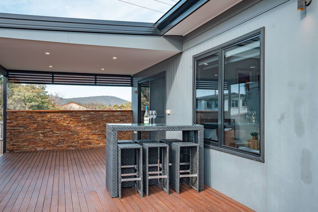 A covered outdoor area with a wooden deck, a stone wall, and a glass table with bar stools.