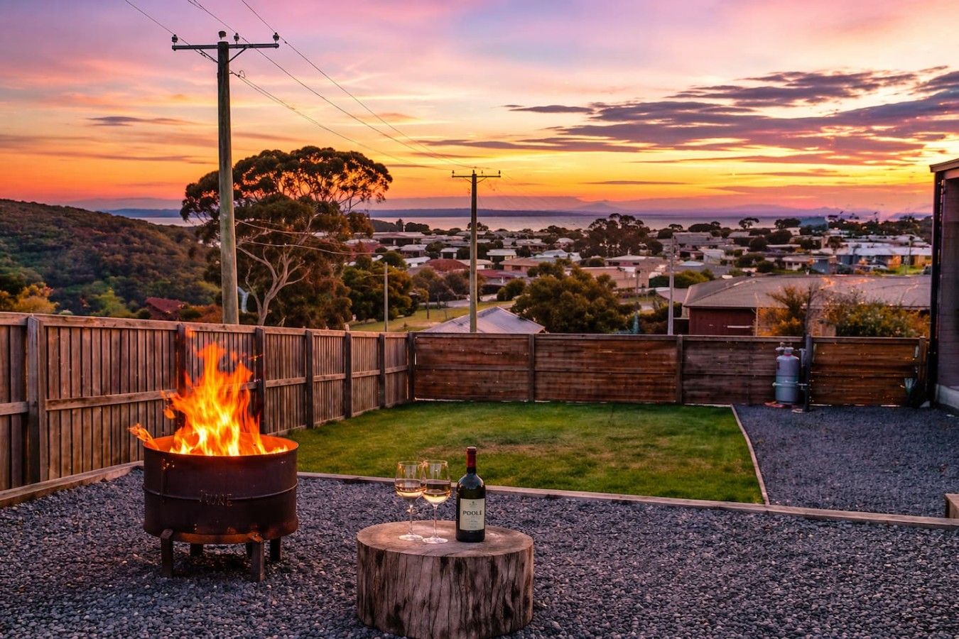 A backyard with a fire pit, a bottle of wine, and a view of a residential area during sunset.
