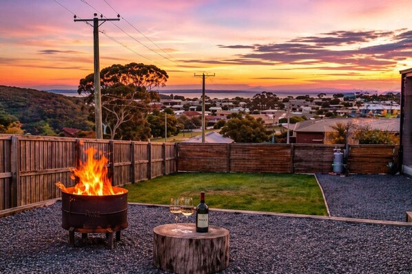 A backyard with a fire pit, a bottle of wine, and a view of a residential area during sunset. - Sorell Hobart 4 Bedroom Family Retreat Near Airport (Sorell)