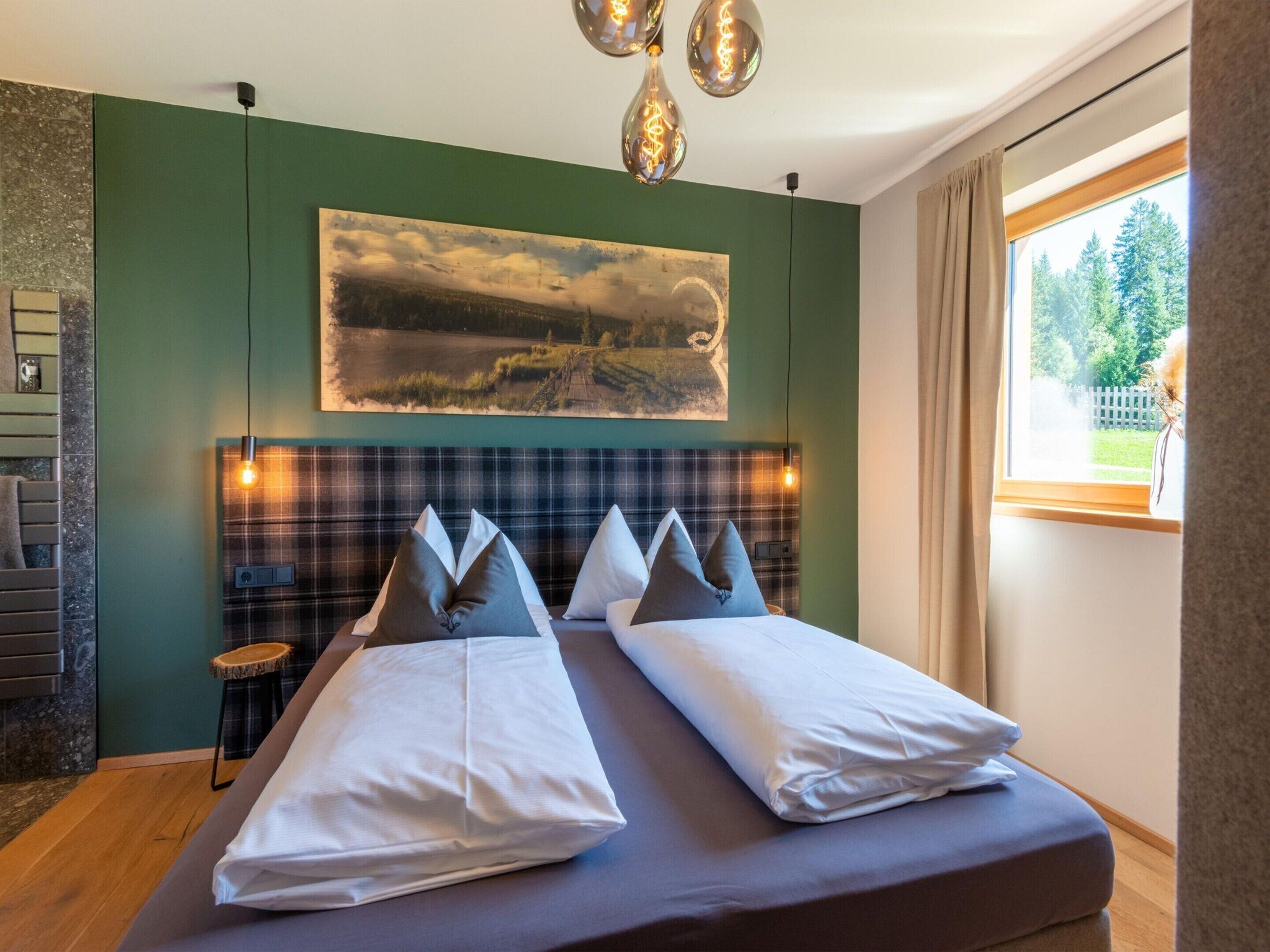 A modern hotel room with a large bed, a headboard with a plaid pattern, a painting on the wall, and a view of trees through the window.