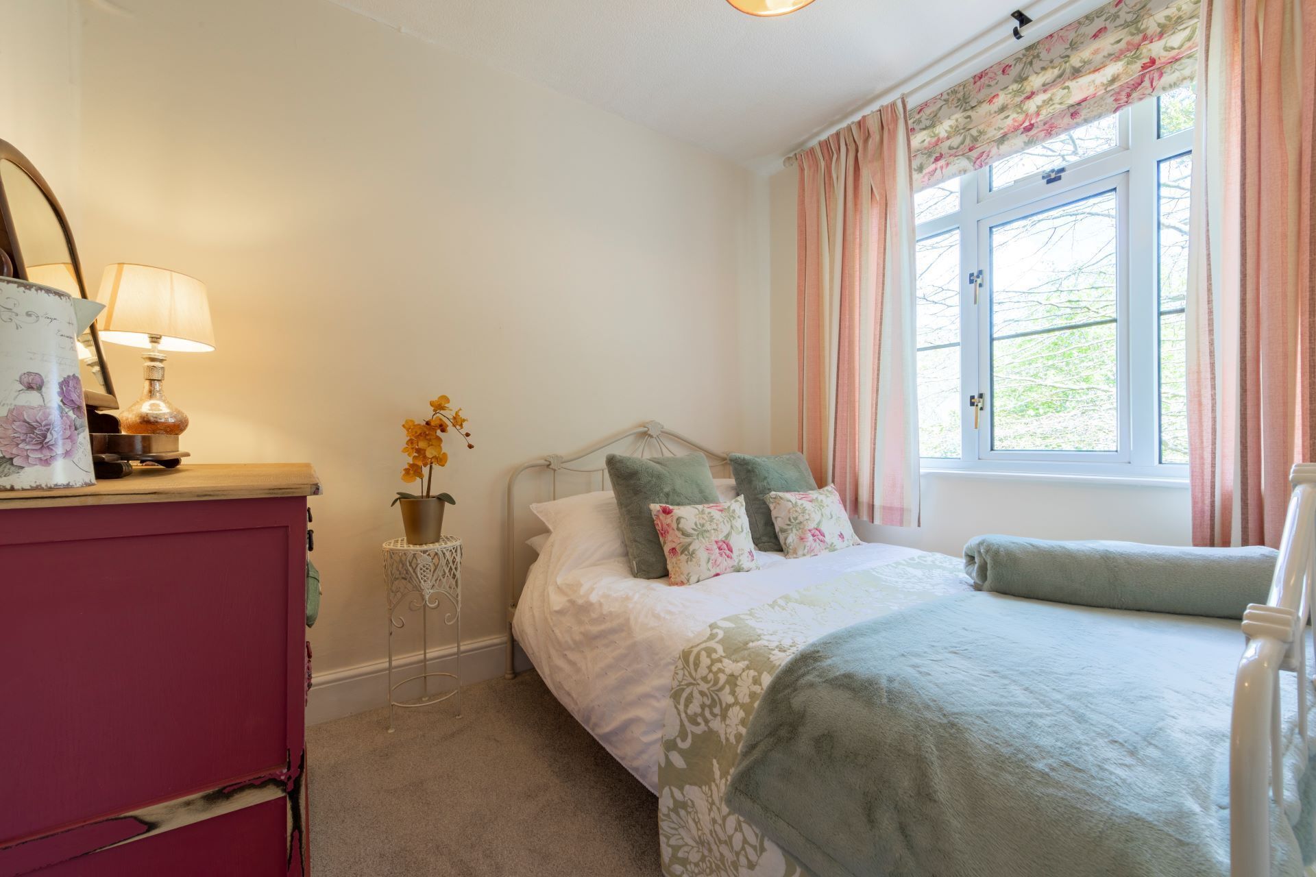 A bedroom with a bed, a nightstand, a lamp, a window with curtains, and a view of greenery outside.