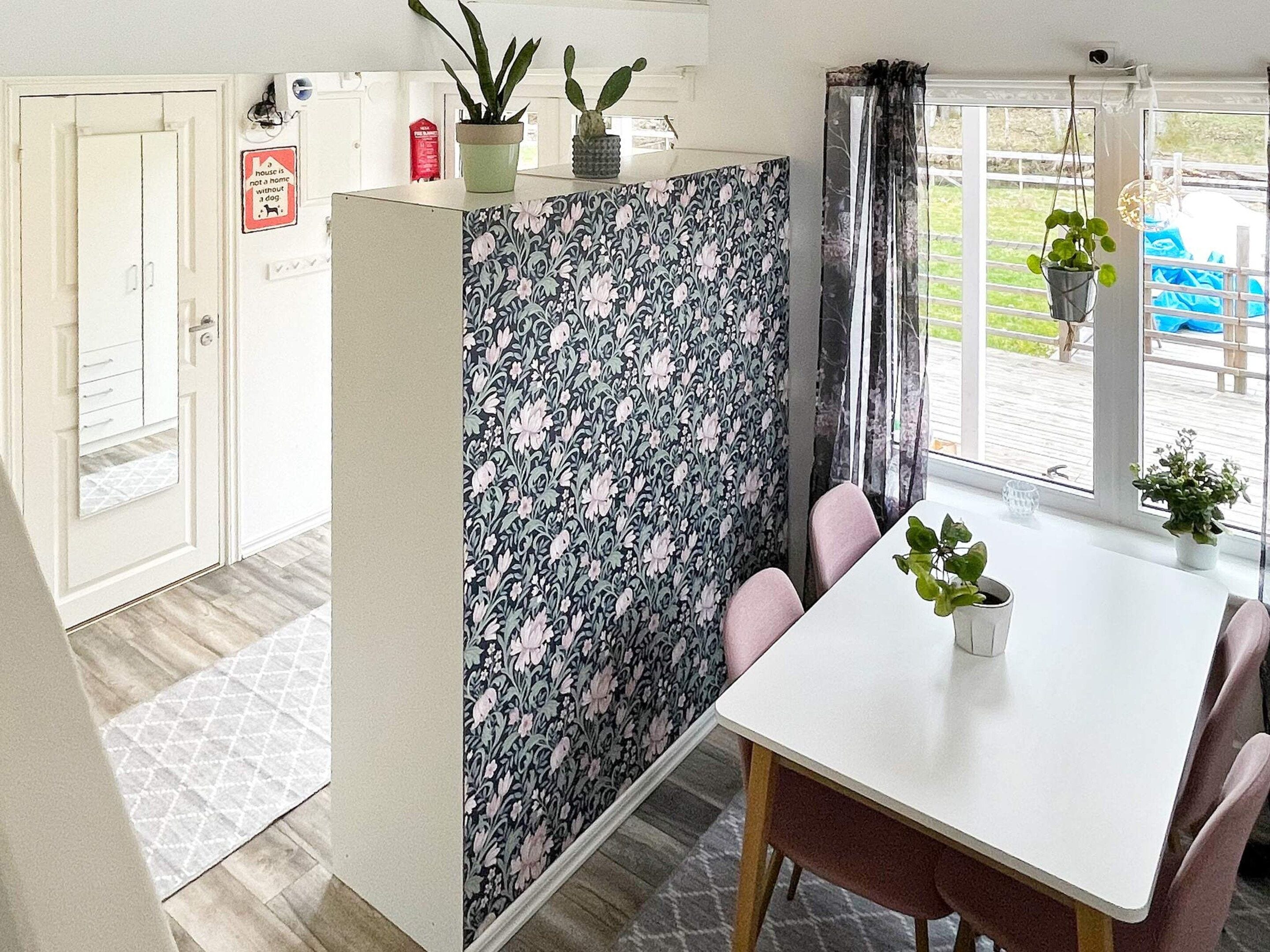 Ein Zimmer mit einem weißen Tisch, rosafarbenen Stühlen, einer trennenden Wand mit Blumenmuster und Blick ins Freie.