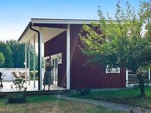 A covered outdoor area with a wooden deck, a red building, and a tree. - 4 Star Holiday Home in Rockneby (Rockneby)