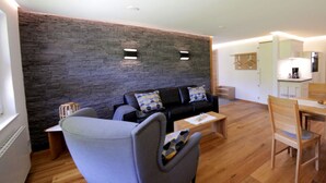 A modern living room with a stone wall, a grey sofa, a wooden dining table, and a kitchen area in the background. - Holiday Apartment Abendgrund (Unterkirnach)