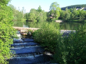 A natural landscape with a small waterfall, dense greenery, and a calm lake.