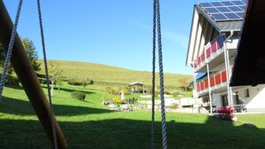 A playground with swings and a netted play area, a building with solar panels, and a grassy landscape with a small structure in the background. - Holiday Apartment Abendgrund (Unterkirnach)