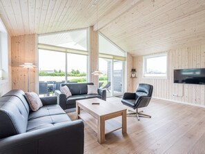 A modern living room with a sofa, armchair, and coffee table, large windows, and a flat-screen TV.