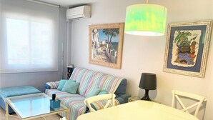 A living room with a striped sofa, a glass coffee table, a white dining table with chairs, and artwork on the walls.