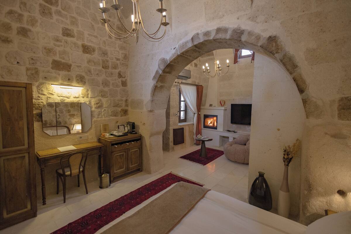A rustic room with stone walls, a wooden desk, a chair, a bed, a fireplace, and a television.