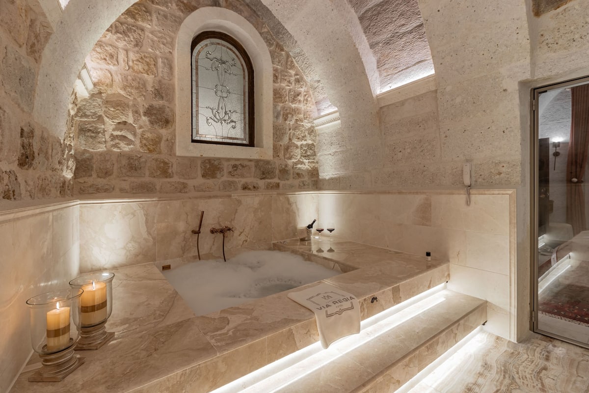 A stone-walled bathroom with a marble bathtub, candles, and a framed picture on the wall.