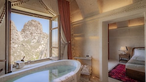A room with a large bathtub, a bed, a nightstand, and a view of a mountainous landscape through a window. - Via Regia Hotel Cappadocia (Nevsehir)