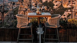 A table with two wicker chairs and a bottle of wine, set against a backdrop of rocky mountains and a clear sky. - Via Regia Hotel Cappadocia (Nevsehir)