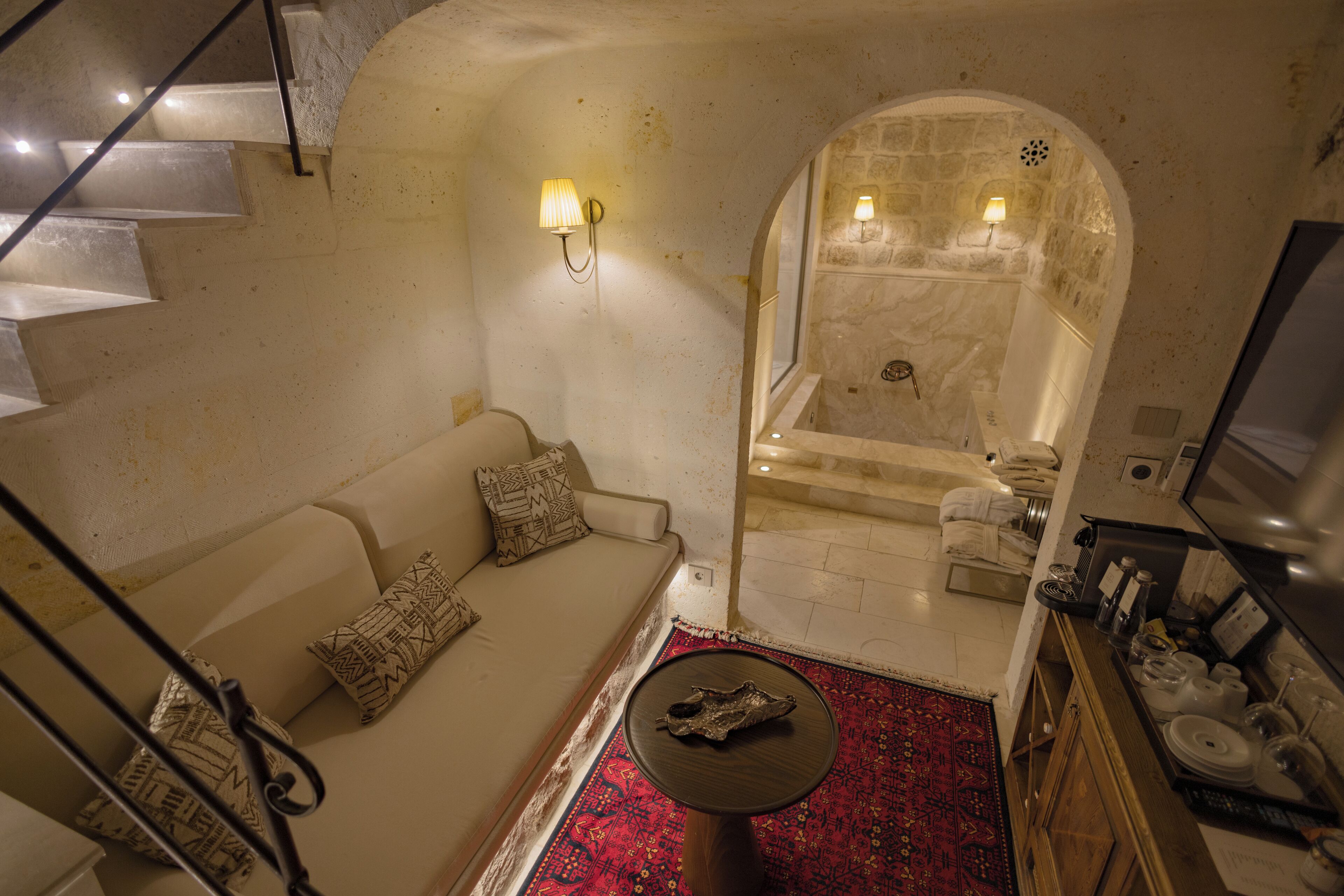 A cozy living area with a sofa, a small round table, a red patterned rug, a stone wall, and a glimpse of a bathroom with a bathtub and a shower.