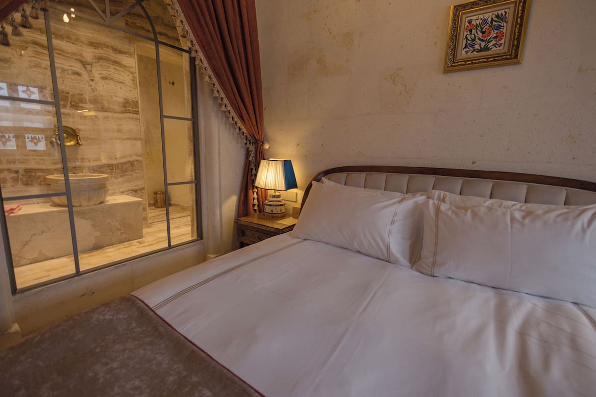 A bedroom with a bed, a nightstand with a lamp, a stone wall, and a window with curtains.