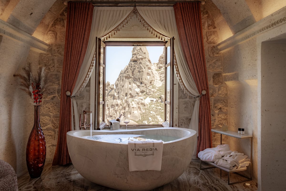 A bathroom with a large freestanding bathtub, a window with curtains, a stone wall, and a vase with dried flowers.