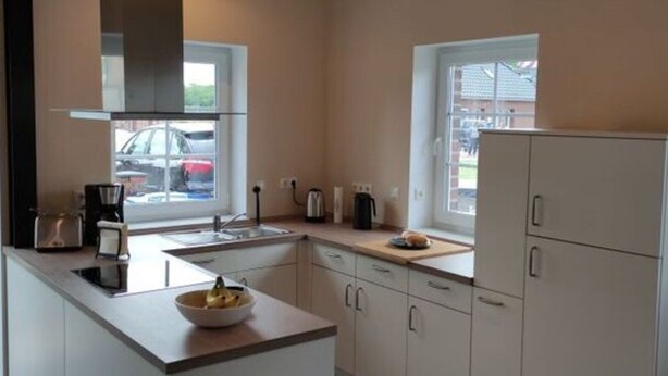 A modern kitchen with white cabinets, a central island, and a large window providing natural light. - Dat Ankerhuus Timmel (Grosefehn)