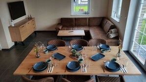 A dining area with a wooden table set for six, surrounded by brown cushioned chairs. A brown leather sofa is against the wall, and a TV is mounted above a wooden cabinet. - Dat Ankerhuus Timmel (Grosefehn)