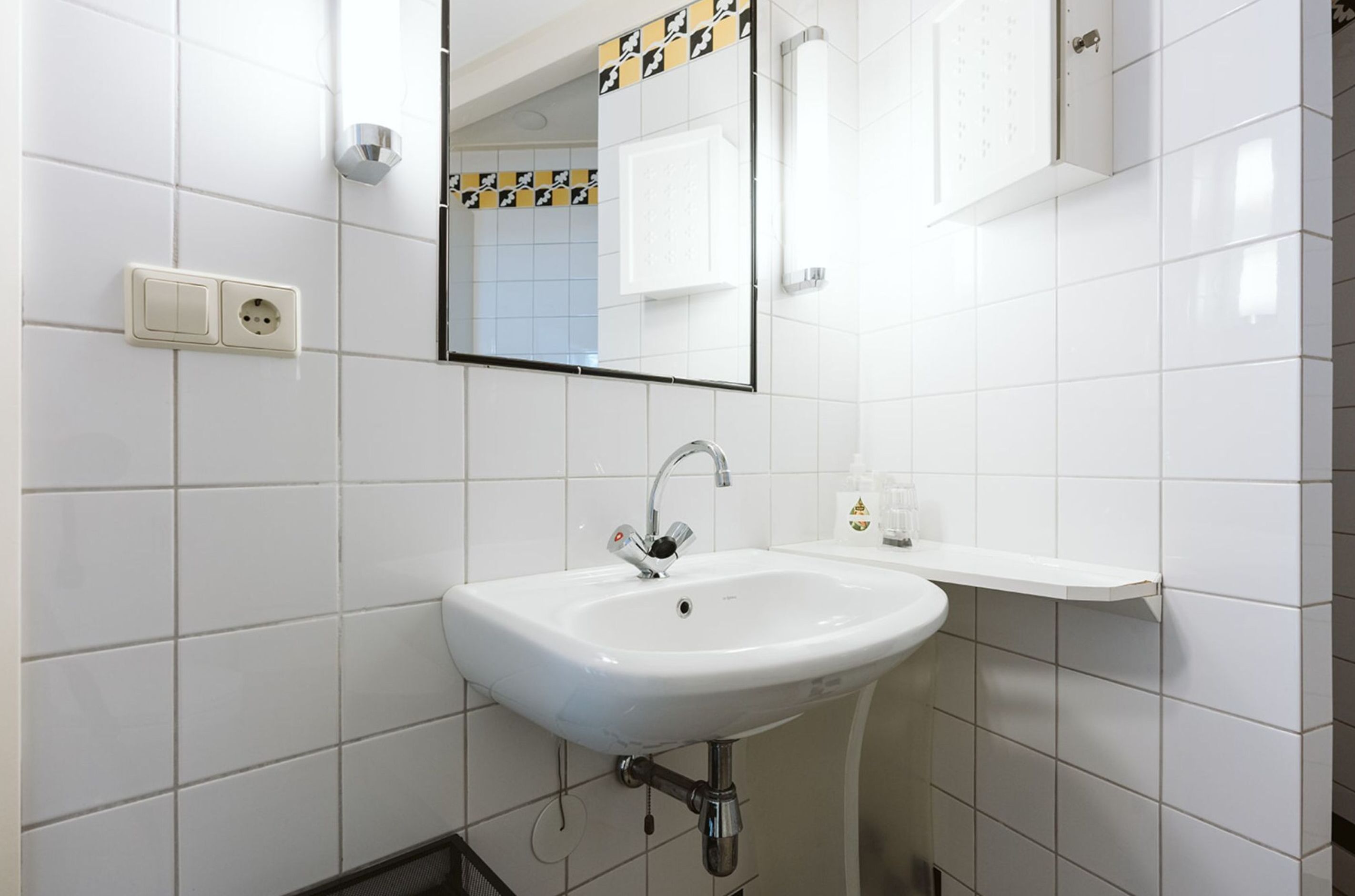 A bathroom with white tiled walls, a white sink with a chrome faucet, a mirror with a black and yellow border, and a white shelf with toiletries.