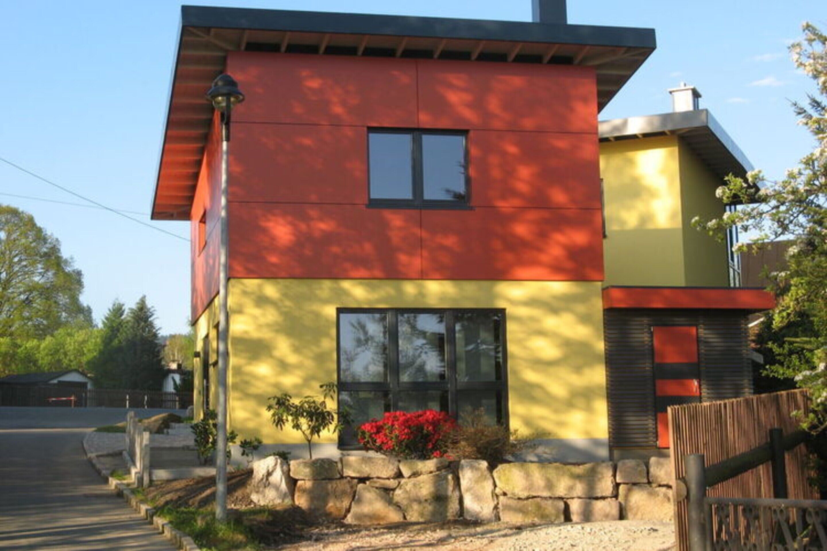 Ein modernes, zweistöckiges Haus mit roter und gelber Fassade, einem Steinfundament und einem Holzzaun.
