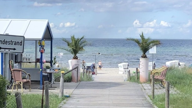 En træpromenade, der fører til et strandområde med en lille bygning med teksten 'indestrand'.